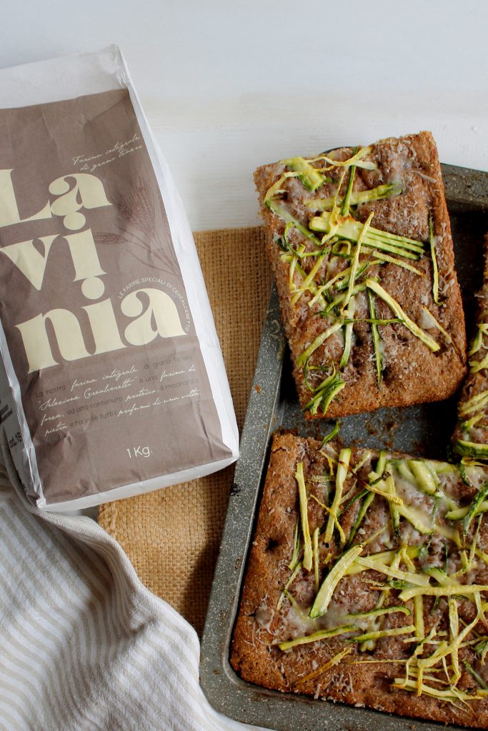Focaccia integrale con zucchine e parmigiano in una teglia grigia, con accanto la confezione della farina Lavinia integrale da 1kg